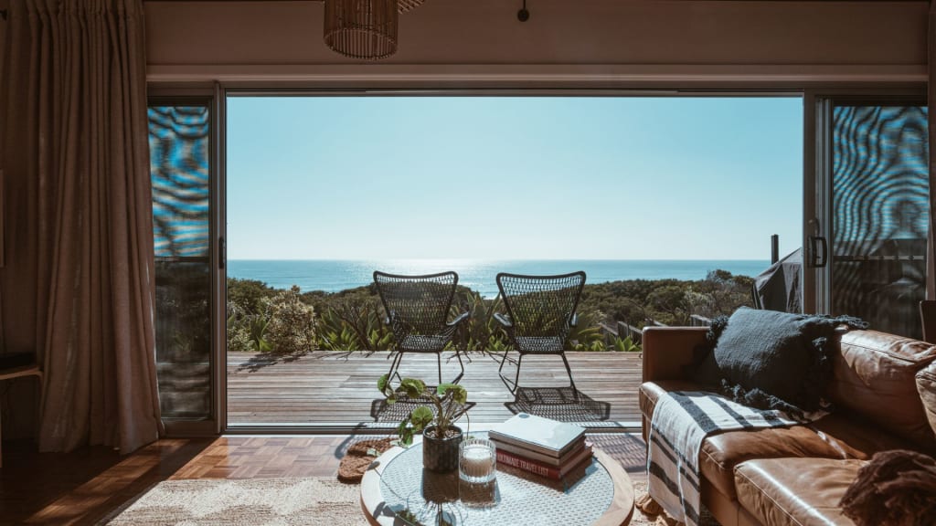 A living room with a round coffee table and a leather sofa opens to a deck with two chairs, overlooking trees and an ocean view under a clear sky.